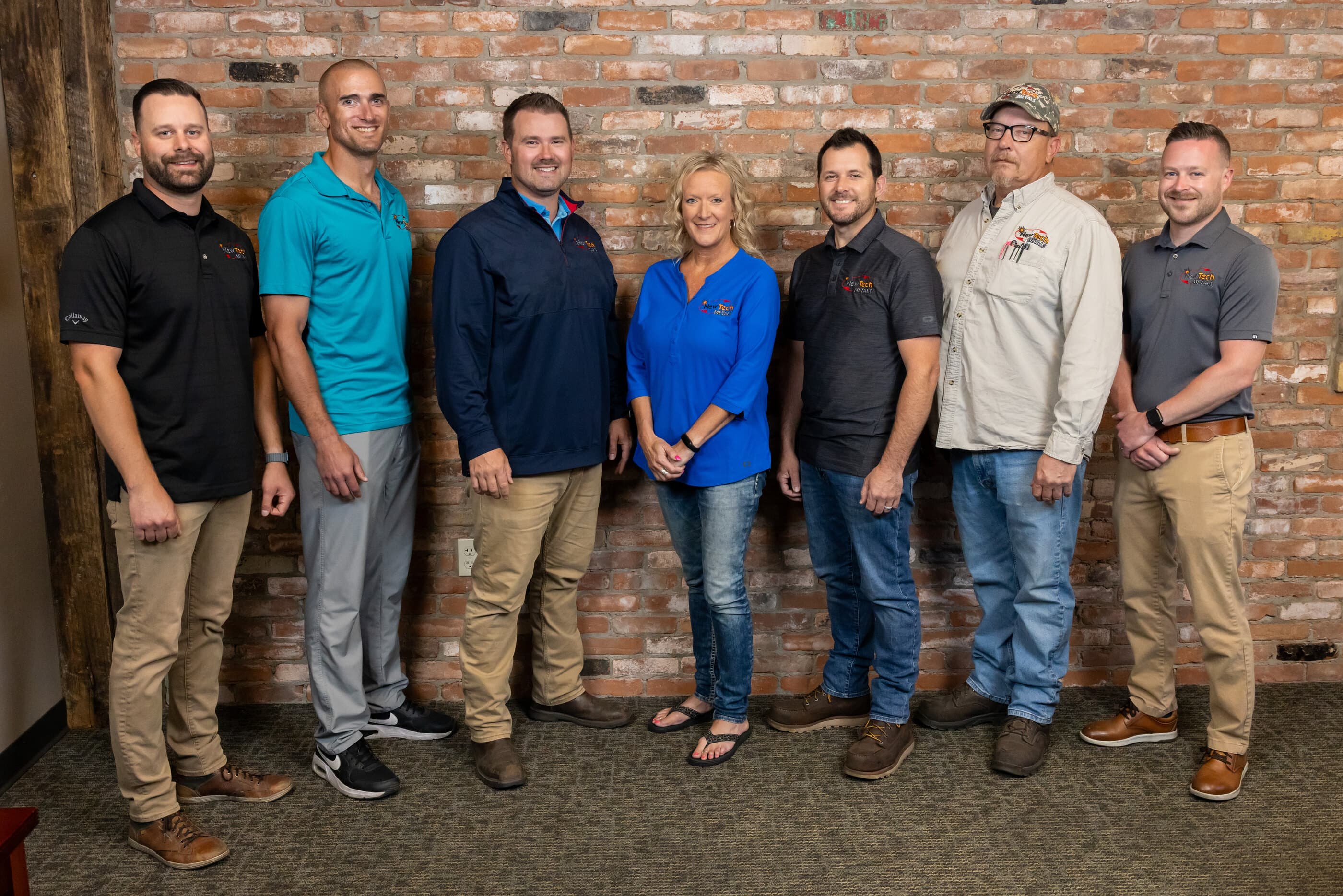 The New Tech Metals management team, seven leaders, including owners Adam, Andy, and Derek Blackman, against the brick wall at the New Franken facility