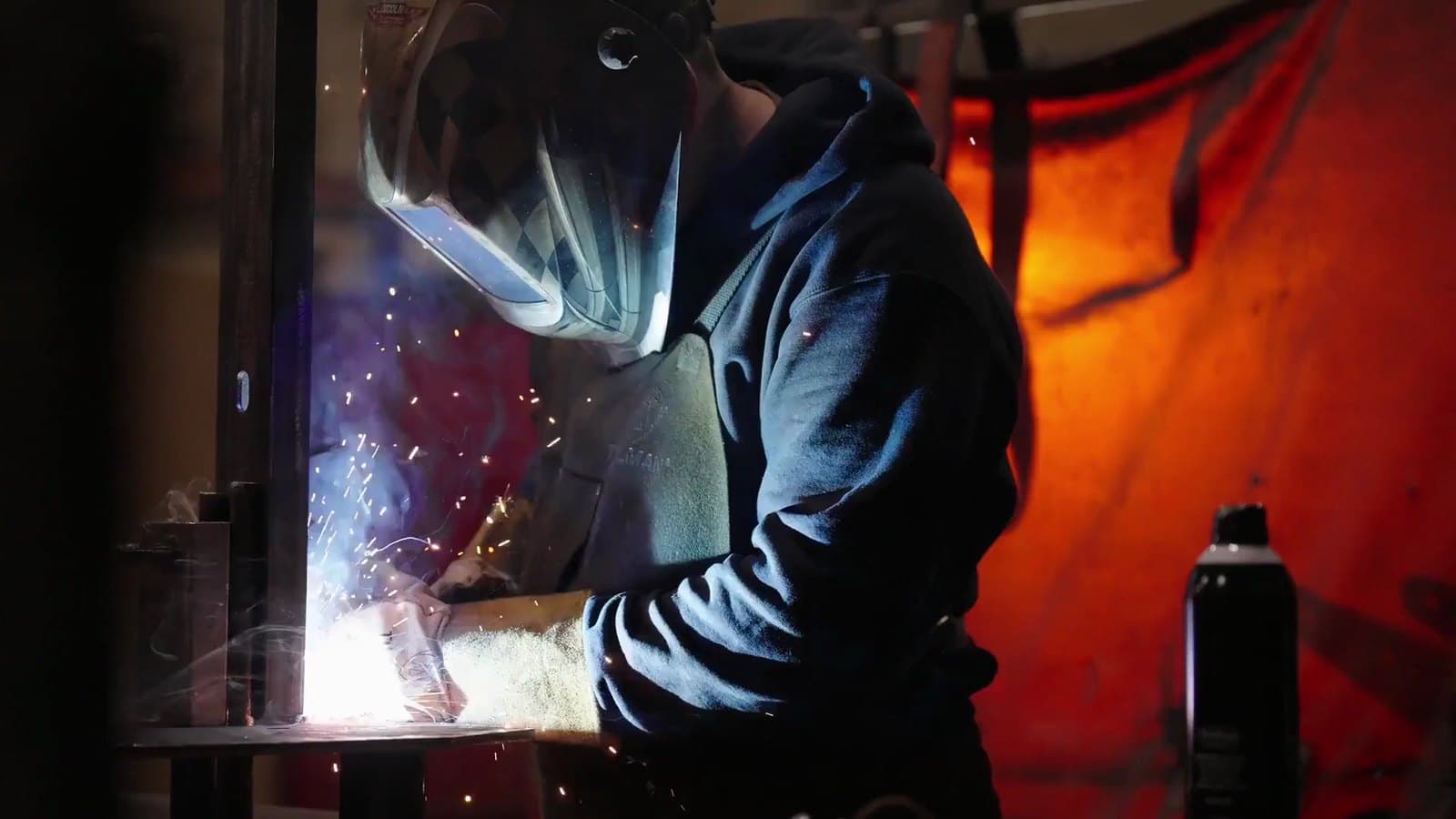AWS-certified welder laying a TIG bead on a steel weldment behind a red welding curtain at New Tech Metals